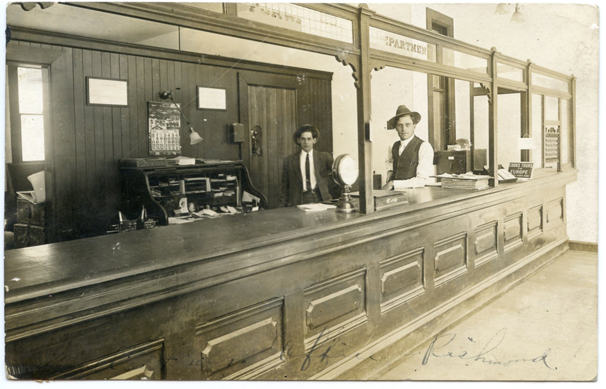 Two men in business attire stand in a room behind a long wooden counter. Furnishings include a roll top desk, wall calendar, and scale on the counter. Handwritten legend reads office Richmond. Image is black and white.