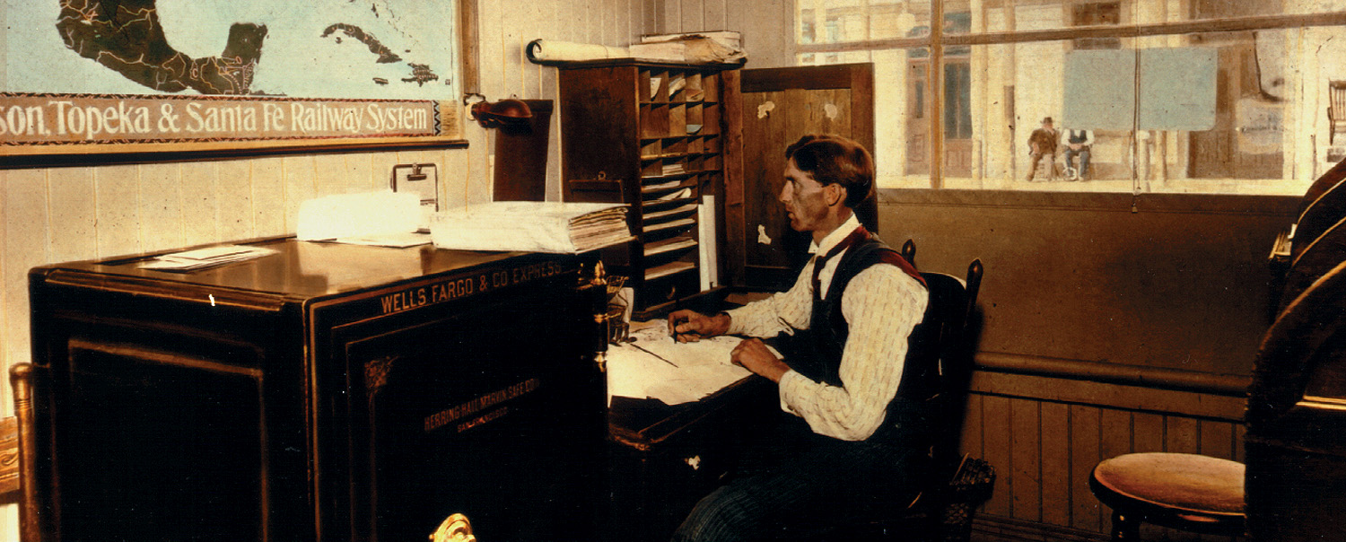 Color picture of a Wells Fargo agent seated at a desk covered in papers next to a safe.
